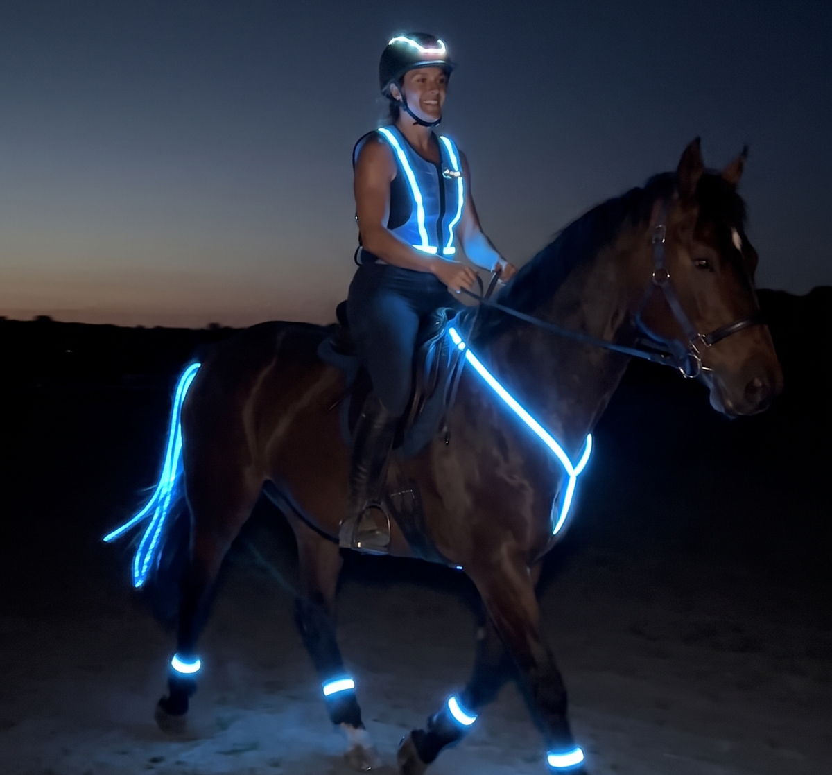 A person riding a horse at night on a sandy path, wearing a reflective safety vest and helmet, with the horse also equipped with glowing reflective bands on its body, legs, and tail.